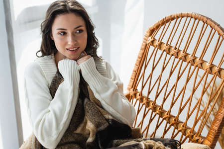 smiling woman sitting in wicker chair under plaid blanket and looking awayの写真素材