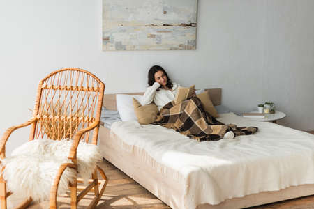 brunette woman reading book in bed under plaid blanket near wicker chairの写真素材
