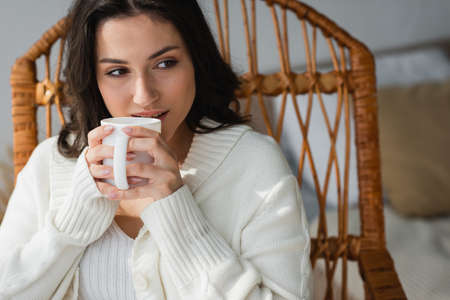 brunette woman in white cardigan sitting in wicker chair and drinking warm tea