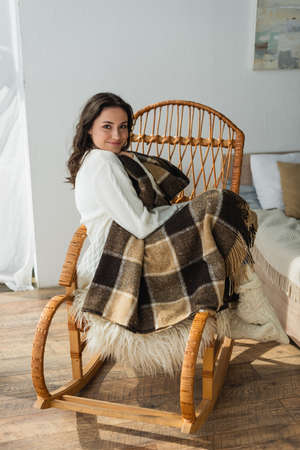 pleased woman looking at camera while sitting in wicker chair under plaid blanketの写真素材
