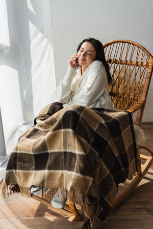 pleased woman sitting in rocking chair under warm blanket and talking on smartphone with closed eyesの写真素材
