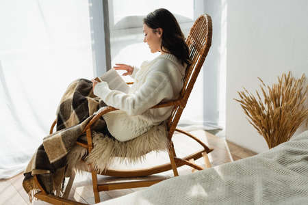 side view of brunette woman reading book in rocking chair under plaid blanketの写真素材