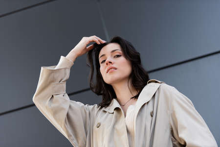 low angle view of brunette woman in beige coat posing with hand near head outdoorsの写真素材