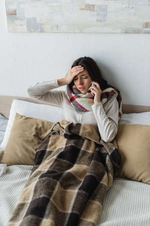 sick woman with closed eyes talking on smartphone while suffering from headache in bedの写真素材