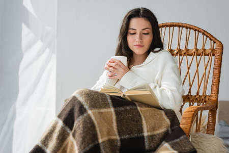 brunette woman sitting in wicker chair with cup of tea and reading novelの写真素材