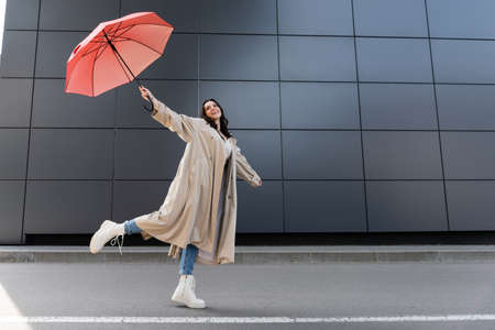 happy woman in beige coat and white boots posing with red umbrella in raised handの写真素材