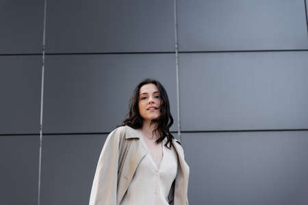 young stylish woman in raincoat looking at camera near gray wallの写真素材