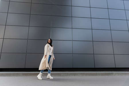 stylish woman in long coat looking back while standing near gray wallの写真素材