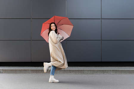 brunette woman in white boots and beige coat looking back under red umbrellaの写真素材
