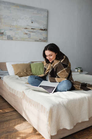 smiling woman sitting on bed under plaid blanket and looking at laptopの写真素材