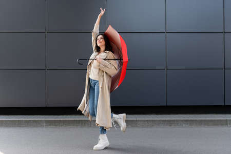 joyful woman in stylish autumn outfit looking up while posing with red umbrella and raised handの写真素材