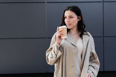 brunette woman in beige raincoat looking away while standing with takeaway drink near gray wallの写真素材