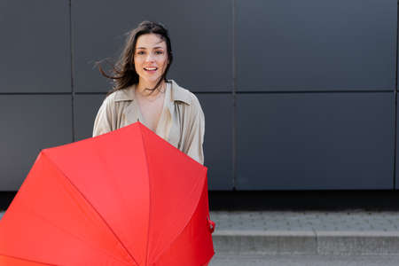 young woman with red umbrella looking at camera while standing on wind near gray wallの写真素材