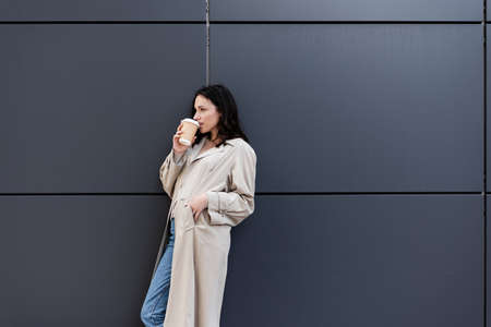 brunette woman drinking coffee while standing near gray wall with hand in pocket of beige coatの写真素材