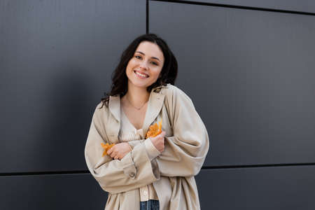young and happy woman in beige coat smiling at camera while standing with yellow leaves near gray wallの写真素材
