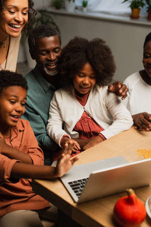 african american boy waving hand near laptop and smiling family during video call on thanksgiving...