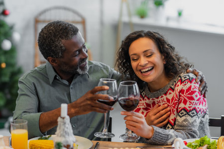 laughing african american woman clinking glasses of red wine with dad during christmas dinnerの写真素材