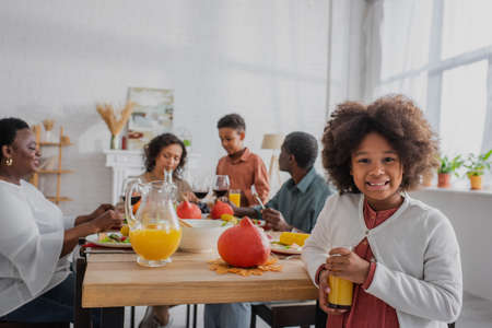 African american kid with orange juice standing near decorated thanksgiving dinner and familyの写真素材