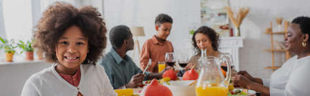 Smiling african american girl standing near blurred family and thanksgiving dinner, bannerの写真素材