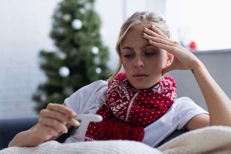 worried woman in warm scarf looking at thermometer near christmas tree on blurred backgroundの写真素材