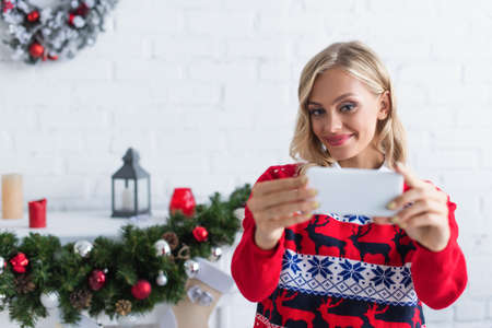 joyful woman in stylish sweater taking selfie on smartphone near blurred christmas garlandの写真素材