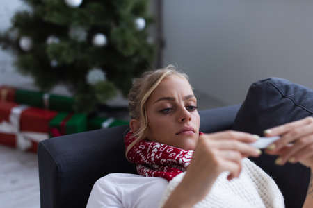 displeased and sick woman on sofa looking at thermometer near christmas tree on blurred backgroundの写真素材