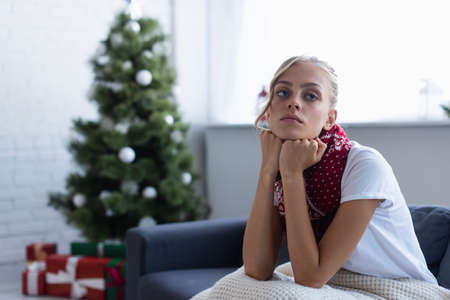 sick and sad woman looking away while sitting on sofa near blurred christmas treeの写真素材