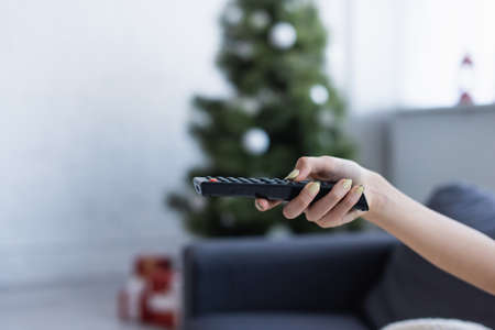 cropped view of female hand with remote controller near blurred christmas treeの写真素材