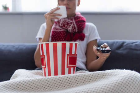 cropped view of sick woman with remote controller and bucket of popcorn watching tv on blurred backgroundの写真素材