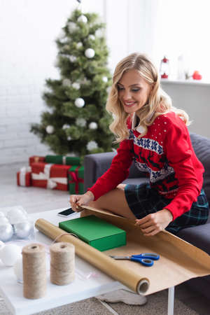happy woman in warm sweater packing gift box into wrapping paper near presents under blurred christmas treeの写真素材