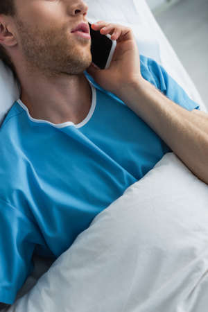 cropped view of man in patient gown talking on smartphone in hospital bedの写真素材