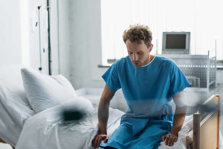 curly man in patient gown sitting on hospital bed