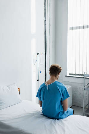 back view of curly man in patient gown sitting on hospital bedの写真素材