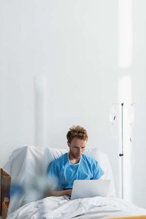 freelancer in patient gown using laptop in hospital bed near drop counterの写真素材