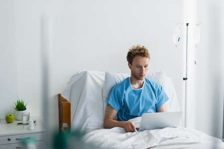 freelancer in patient gown using laptop in hospital bedの写真素材