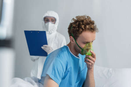 sick patient breathing in oxygen mask near blurred doctor in personal protective equipment holding clipboardの写真素材