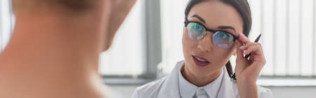 brunette doctor with pen adjusting glasses while looking at blurred patient in hospital, bannerの写真素材