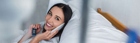smiling woman with oximeter on finger talking on smartphone while lying on hospital bed, bannerの写真素材