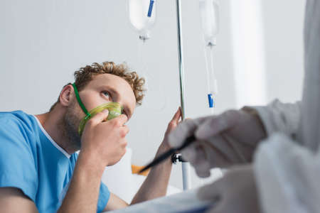 patient in oxygen mask looking at blurred doctor with clipboard and penの写真素材