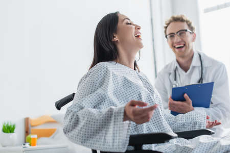 cheerful woman in wheelchair laughing near blurred doctorの写真素材
