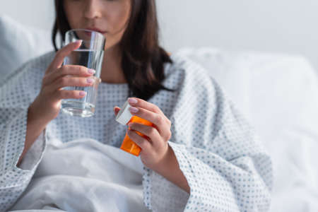 cropped view of sick woman holding bottle with medication while taking pill in hospital