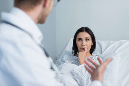 brunette woman in patient gown looking at blurred doctor in hospitalの写真素材