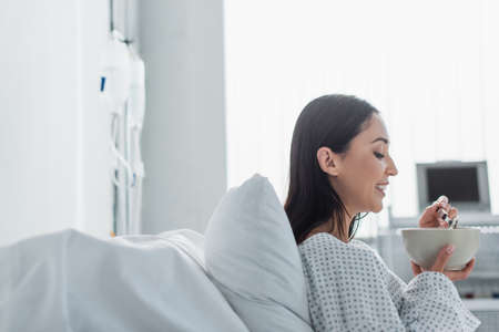side view of cheerful woman holding bowl with breakfast in hospitalの写真素材