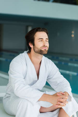 Young man in white bathrobe sitting on deck chair in spa centerの写真素材