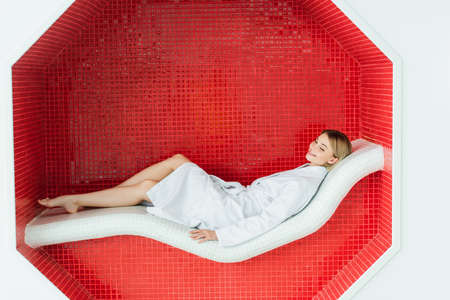 Cheerful woman in white bathrobe lying on lounge chair in spa centerの写真素材