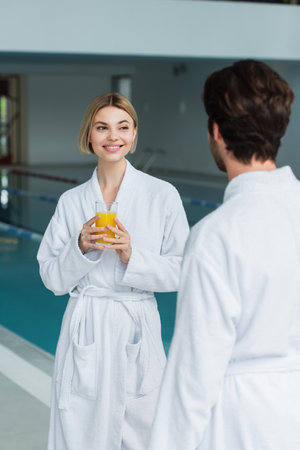 Cheerful woman holding glass of orange juice near blurred boyfriend in white bathrobe in spa centerの写真素材