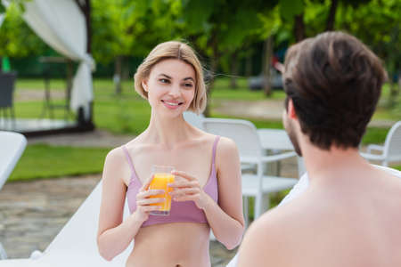 Smiling woman in swimsuit holding glass of orange juice near blurred boyfriend on resortの写真素材