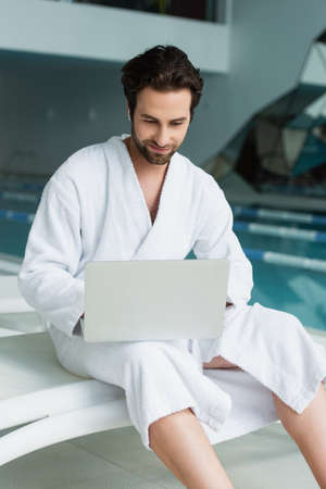 Young man in bathrobe and earphone using laptop in spa centerの写真素材