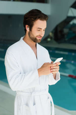 Young man in wireless earphone and bathrobe using smartphone in spa centerの写真素材