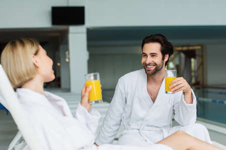 Smiling man in bathrobe holding glass of orange juice near blurred girlfriend in spa centerの写真素材